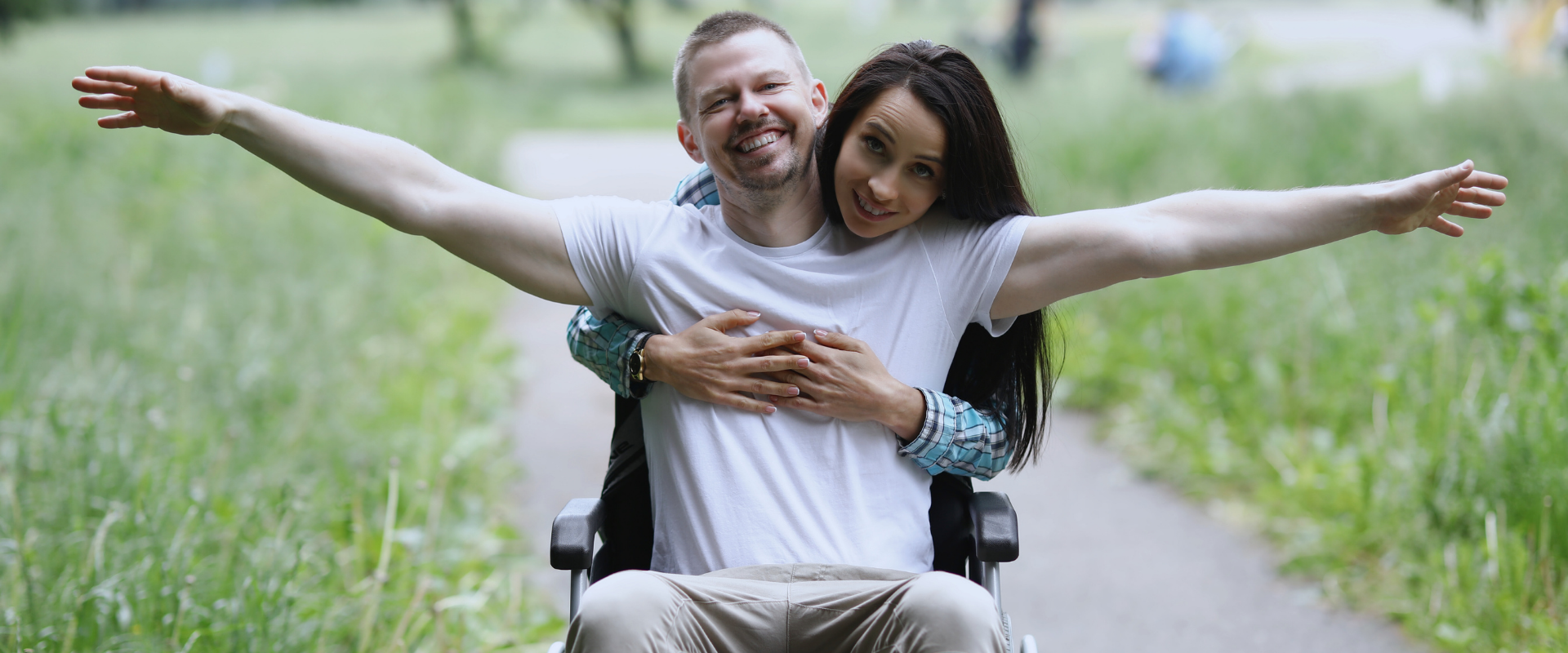 A joyful couple outdoors - a woman hugging a man in a wheelchair, both with arms spread wide and smiling