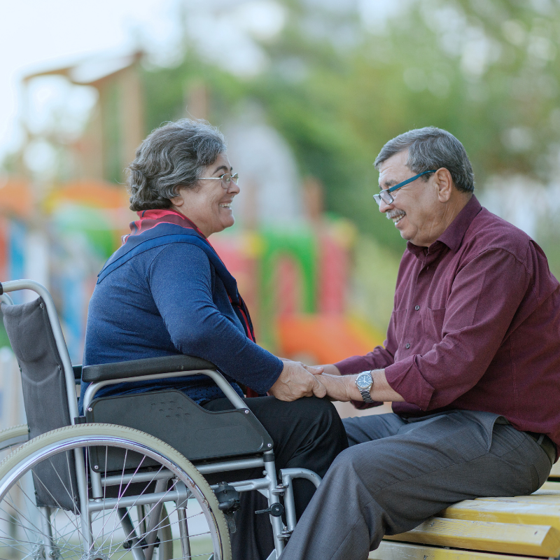 A couple sharing a warm moment outdoors, one partner in a wheelchair holding hands with the other
